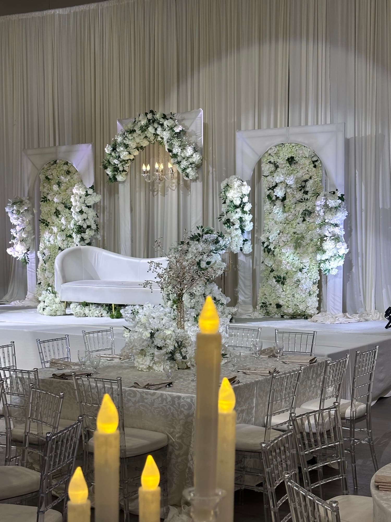 White floral arch backdrop and head table set with clear chiavari chairs for a three-hundred-guest event at Palacio Event Centre